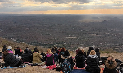Grupo de personas sentadas de espaldas en una colina, observando un vasto paisaje boscoso y montañoso con el cielo del atardecer en el horizonte.
