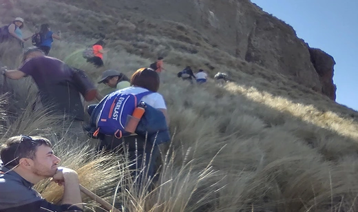 Grupo de senderistas ascendiendo una ladera empinada y cubierta de hierba alta en una montaña, bajo un cielo despejado.