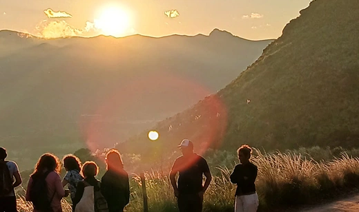 Grupo de personas de pie en una colina, observando un atardecer radiante sobre un paisaje montañoso.