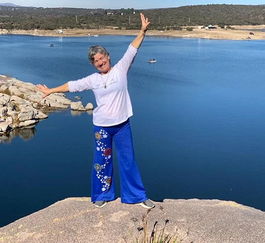 Mujer sonriente de pie en una roca junto a un gran lago azul, con los brazos extendidos y una pose de alegría.